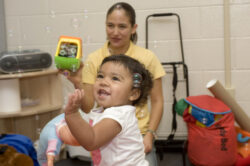 A smiling toddler plays with bubbles indoors while an adult sits in the background holding a bubble maker. Colorful bags and toys are scattered around the room.
