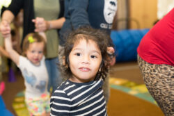 A young child with long hair and a striped shirt looks at the camera, while other children and adults are seen playing and holding hands in a brightly lit indoor space.