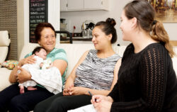Three women sit on a couch; one holds a baby. They are smiling and talking, with a notepad and pen visible, suggesting a friendly or consultative discussion in a cozy home setting.
