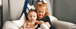 Baby and Toddler sit next to one another in a chair, playing and smiling.