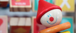 Primer plano de un payaso de juguete de madera con un sombrero rojo, cara sonriente y aros de colores, con bloques con letras desenfocados al fondo.