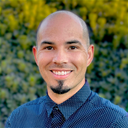 Smiling man with a shaved head and goatee, wearing a navy blue patterned button-up shirt, standing outdoors with greenery in the blurred background.