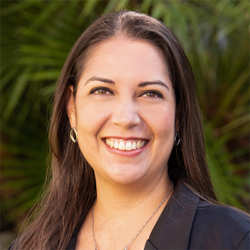 Woman with long brown hair smiling, wearing a black top and silver earrings, standing outdoors with green leafy plants in the blurred background.
