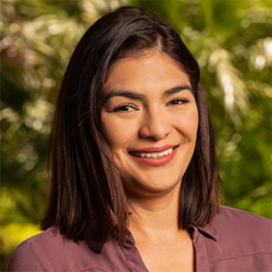 A woman with straight dark hair, wearing a mauve blouse, smiles at the camera. The background is filled with green, sunlit foliage.