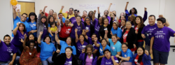 A large, diverse group of adults and children pose together in a room, many raising their fists and smiling. Most are wearing matching blue, purple, or red shirts, suggesting a sense of unity or participation in an event.