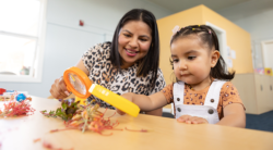 An adult and a young girl examine plants with a magnifying glass at a table in a bright classroom. The woman smiles as the child focuses on the leaves.