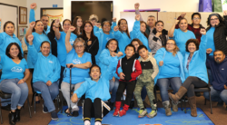 A diverse group of adults and children, many in matching blue shirts, smile and raise fists in celebration while posing together in a colorful indoor setting.