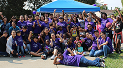 Un numeroso grupo de personas, la mayoría con camisetas moradas, posa al aire libre bajo un toldo azul; muchas de ellas levantan el puño y sonríen, mientras que los niños están sentados y de pie delante.