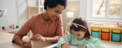 A woman smiles while sitting beside a young girl who is drawing with colored pencils at a wooden table in a bright, organized room.