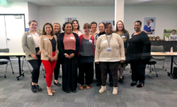 Doce mujeres están juntas en una sala de reuniones, sonriendo para una foto de grupo. Visten ropa informal de oficina. Detrás de ellas hay mesas y sillas dispuestas en fila.
