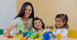 A woman and two young girls are smiling at a table covered with colorful plastic building toys, with a plain beige background.