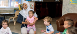 A woman in a hijab sits with a toddler on her lap, surrounded by four young children. They are indoors, sitting on the floor, and appear to be engaged in an activity together.