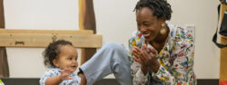 A smiling adult sits on the floor, clapping and interacting joyfully with a happy baby who is reaching out with both hands. They are in a warmly lit indoor space.