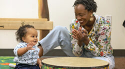 A smiling woman claps while sitting beside a happy baby who is reaching toward a large drum in a colorful indoor setting.