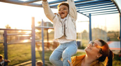 A smiling child hangs from playground bars while an adult supports them from below. The sun is setting, casting a warm glow on the scene.