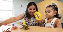 A woman and a young girl sit at a table. The girl uses a yellow magnifying glass to examine colorful flowers, while the woman smiles and reaches for another flower.