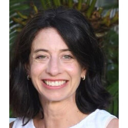 A woman with shoulder-length dark hair and a white top smiles outdoors, with green plants visible in the background.
