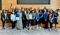A diverse group of adults, some holding certificates, stand together and smile inside a modern meeting room with wood paneling and an American flag in the background.