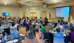 A large group of people sits at round tables in a bright conference room, listening to a woman speaking at a podium with a projected screen behind her. The tables have food, drinks, and books.
