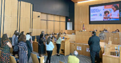 People stand and present to officials in a wood-paneled council chamber. A large screen displays Investing in Childrens Most Critical Years with a smiling woman and child. An American flag and seal are visible.
