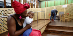 A woman wearing a red headwrap kisses a baby on the cheek while sitting indoors. In the background, another adult and child are near a play area with small tables and chairs.