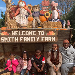 A family of five sits in front of a Welcome to Smith Family Farm sign, surrounded by hay, pumpkins, and autumn decorations with scarecrows and sunflowers.