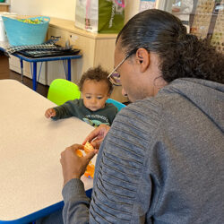 An adult peels an orange at a table while a young child sits across, watching. The setting appears to be a classroom or daycare with toys and supplies in the background.