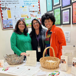 Three smiling people stand behind a table with informational materials, pens, and baskets, in front of a wall covered with colorful flyers and posters promoting community resources.