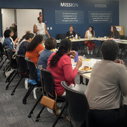 A group of women sit around tables in a meeting room, eating and listening to a woman standing and speaking. Mission and vision statements are displayed on the wall behind them.