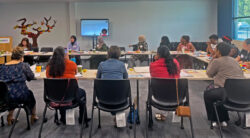 Un grupo de personas está sentado alrededor de unas mesas grandes en una sala de reuniones, escuchando una presentación. En la parte delantera hay una pantalla en la que se proyectan diapositivas, y sobre las mesas hay materiales de bellas artes y hojas de papel.