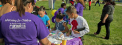 Children and adults gather outdoors at a community event. Kids work on crafts at a table, while a woman in a purple shirt with advocacy text stands nearby. Bright balloons and other people are visible in the background.