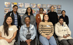 A group of nine women smiling and posing indoors in front of a wall decorated with colorful photos and motivational signs. Four are seated in front, while five stand behind them.