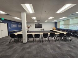 A modern conference room with a U-shaped arrangement of tables and black chairs, a wall-mounted screen, and large windows with blinds. Motivational text is on one wall. The room has gray carpeting and overhead lights.