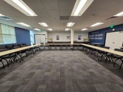 Large meeting room with U-shaped tables and black chairs, blue and white walls, carpeted floor, overhead lights, and posters on the walls. No people are present.