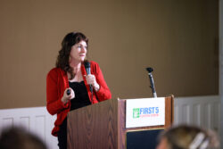 A woman in a red cardigan speaks into a microphone at a podium labeled FIRST 5 Contra Costa during a presentation or event.