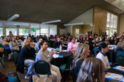 A large group of people sit at round tables in a spacious, well-lit room, attentively listening to a speaker out of view. Some have notebooks, drinks, and papers in front of them.