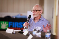 An older man with glasses and a checkered shirt speaks into a microphone at a panel, gesturing with his hand. Three water bottles and a name tag are visible on the table in front of him.