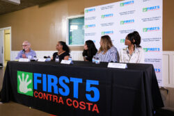 Five people sit at a panel table with a First 5 Contra Costa banner, speaking at an event. The backdrop features logos and phrases about investing in childrens critical years. Bottled water and microphones are on the table.