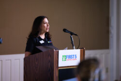 A woman speaks at a podium with a microphone. The podium has a First 5 Contra Costa sign. She holds a folder and wears a name tag. The background is plain and softly lit.