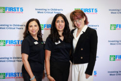Three women stand together smiling in front of a step-and-repeat banner displaying First 5 Contra Costa and Investing in Childrens Most Critical Years. Two women wear black tops; one wears a black blazer over a white blouse.