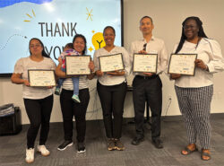 Five people stand indoors, smiling and holding certificates. A screen behind them displays Thank You with decorative elements. One person holds a small child. Everyone is dressed in business casual attire.