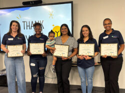 Five women stand in a row, each holding a certificate. One woman holds a small child. Behind them is a screen displaying Thank You! with yellow and blue decorations. All are smiling and appear to be celebrating.
