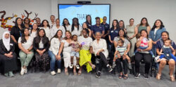 A diverse group of adults and a few young children pose together, smiling, in a brightly lit room. A screen behind them displays THANK YOU above a sun illustration.