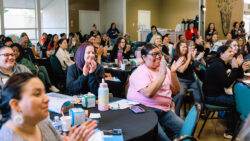 A diverse group of people sit at round tables in a bright room, smiling and clapping, while attending a lively indoor event or seminar.
