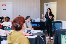 A woman stands and speaks into a microphone at an indoor event, addressing seated attendees. A table with a sign reading First 5 Contra Costa is visible behind her.