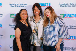 Three women stand together smiling in front of a backdrop that reads FIRST 5 Contra Costa and Investing in Childrens Most Critical Years..