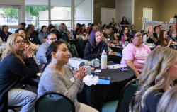 A diverse group of people sit at round tables in a bright room, attentively listening to a speaker. Some are taking notes, and various drinks and papers are on the tables.