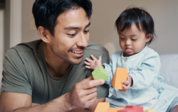 A man smiles while playing with a toddler who is holding colorful blocks. They are lying on a bed, enjoying a playful moment together.