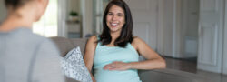 A pregnant woman sits on a couch, smiling and holding her belly while talking to another person whose back is facing the camera.