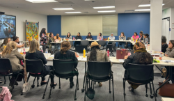 A group of women sit around a large U-shaped table in a conference room, listening to a speaker. Drinks and papers are on the tables, and a screen shows a video call in the corner.